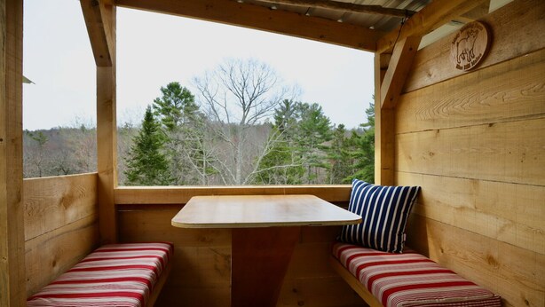 A wooden cabin with a bench and a table, a wooden wall with a logo, and a view of trees outside. - Tiny Cabin on Blue Hill Farm (Blue Hill)
