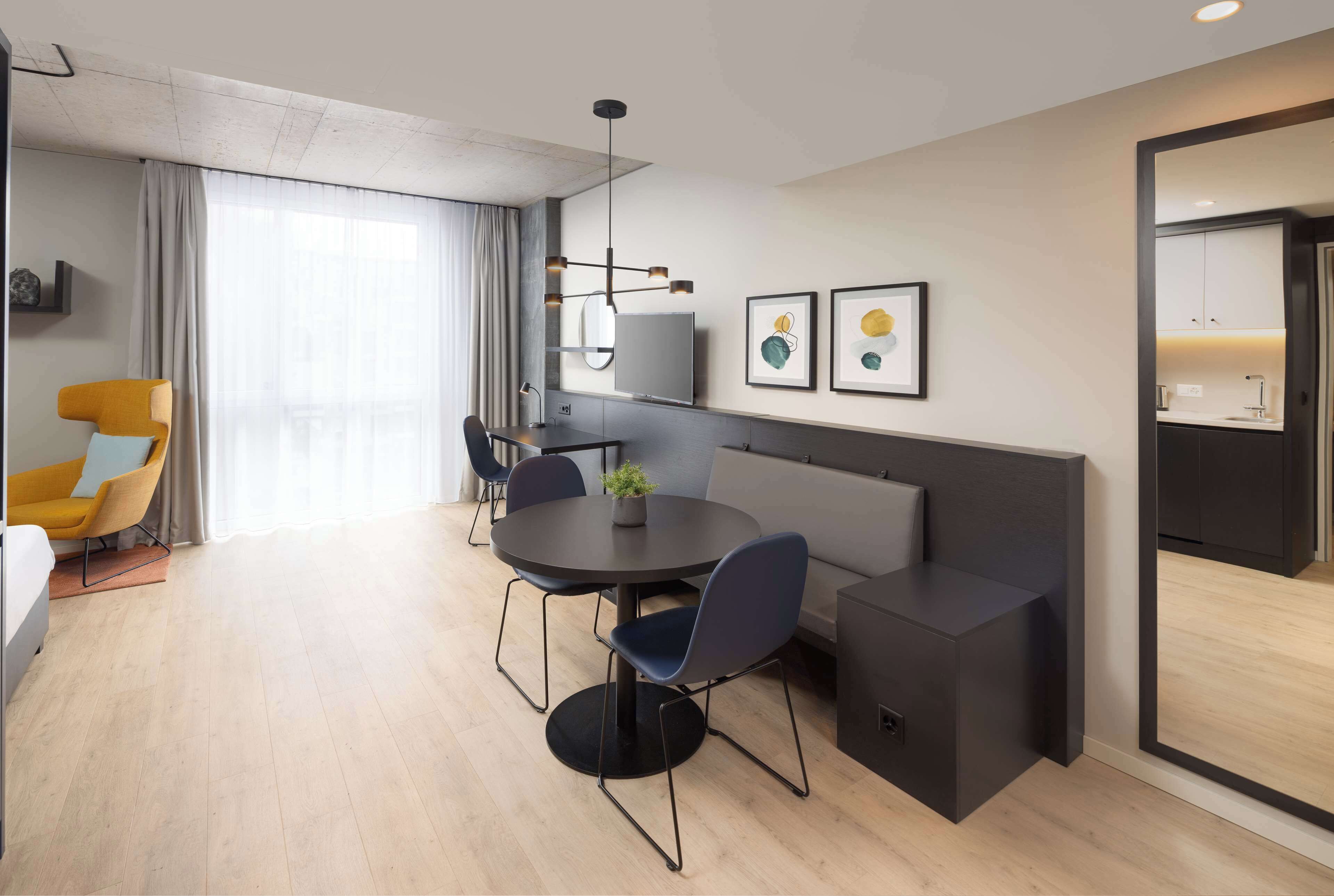 A modern hotel room with a round dining table, two blue chairs, a sofa, a TV, and artwork on the wall.