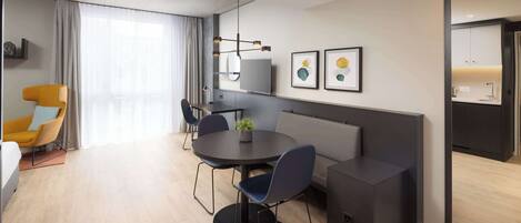 A modern hotel room with a round dining table, two blue chairs, a sofa, a TV, and artwork on the wall.
