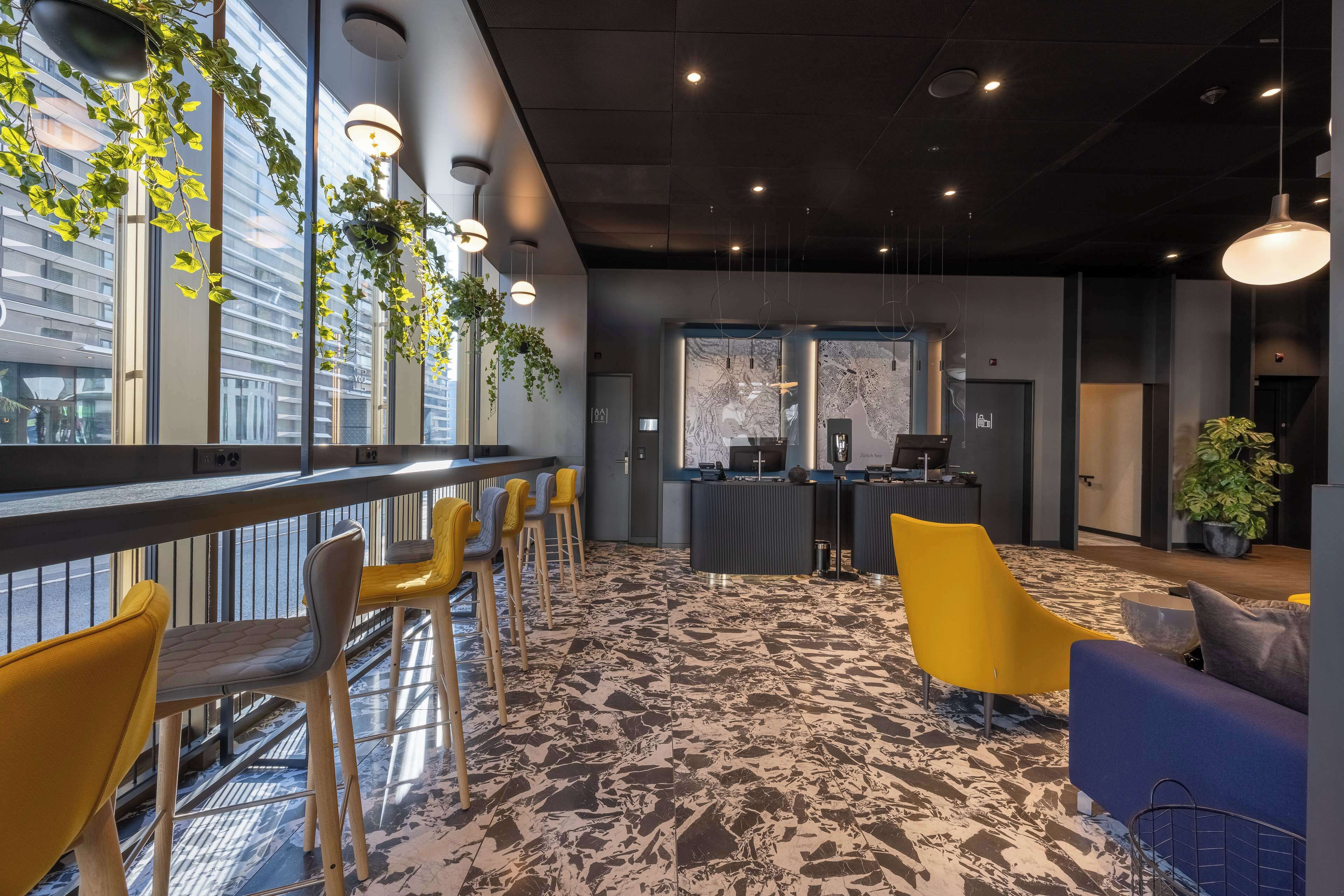 Un intérieur moderne avec un espace bar, des chaises jaunes, un comptoir d’accueil et de grandes fenêtres ornées de plantes.