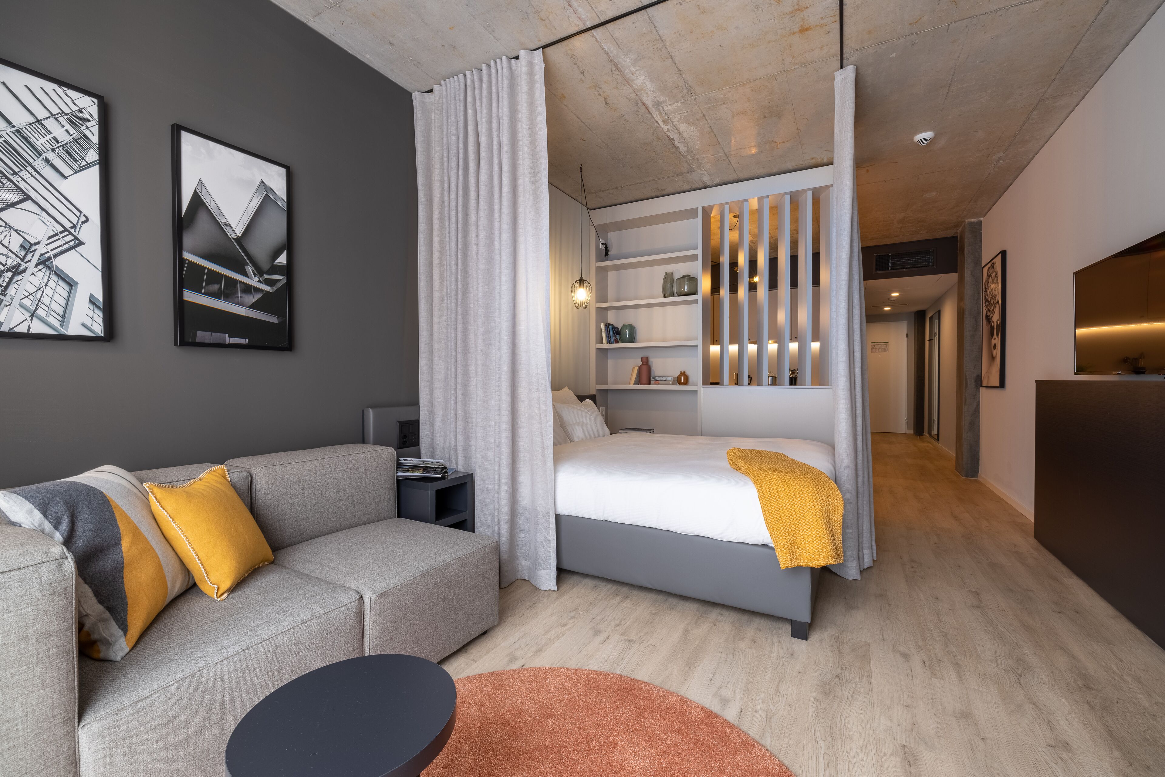 Une chambre d’hôtel moderne avec un canapé gris, un lit doté d’une tête de lit blanche, une petite table et une grande fenêtre avec des rideaux.