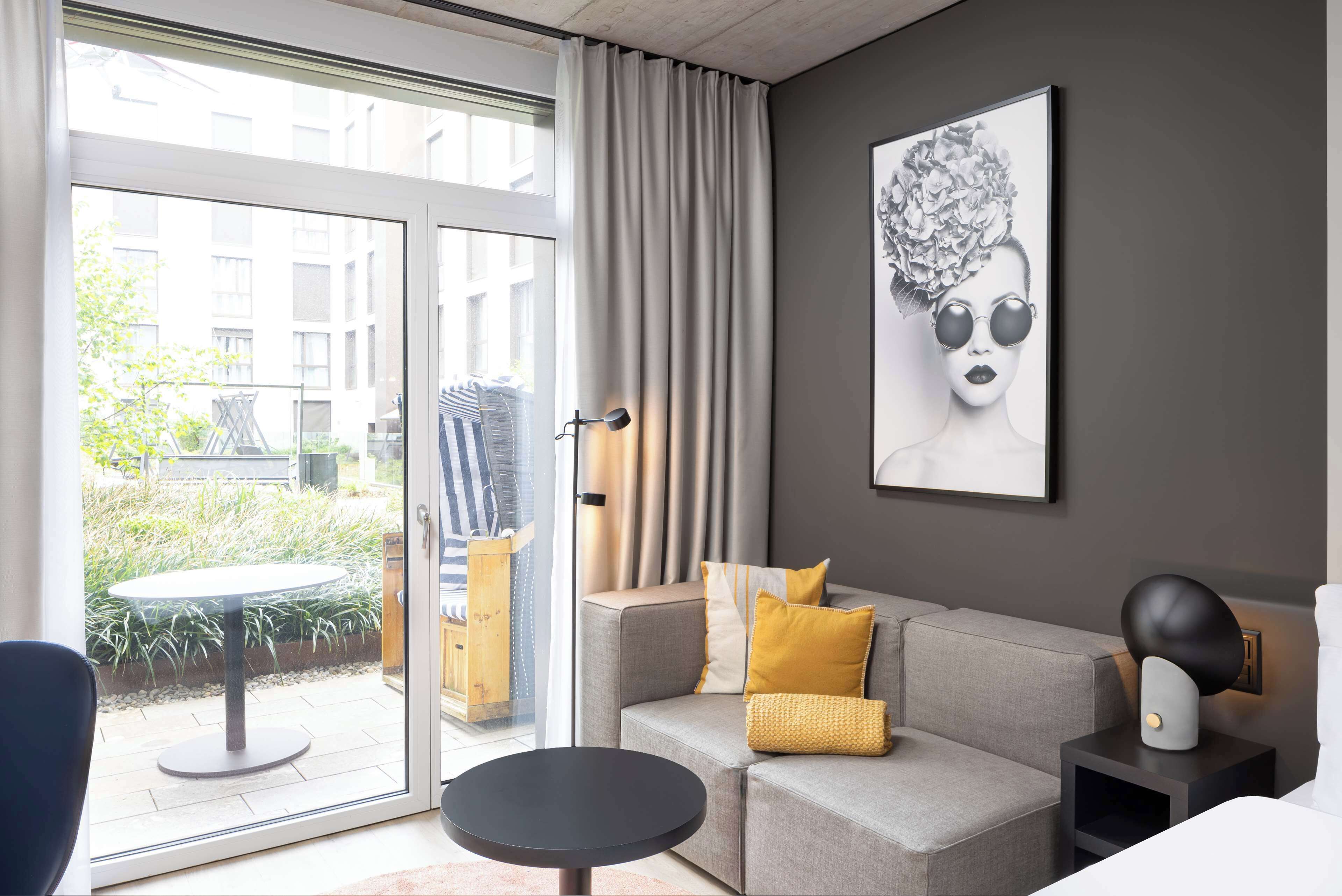 Una sala de estar moderna con un sofá gris, cojines amarillos, una mesa redonda negra y un amplio ventanal con cortinas.