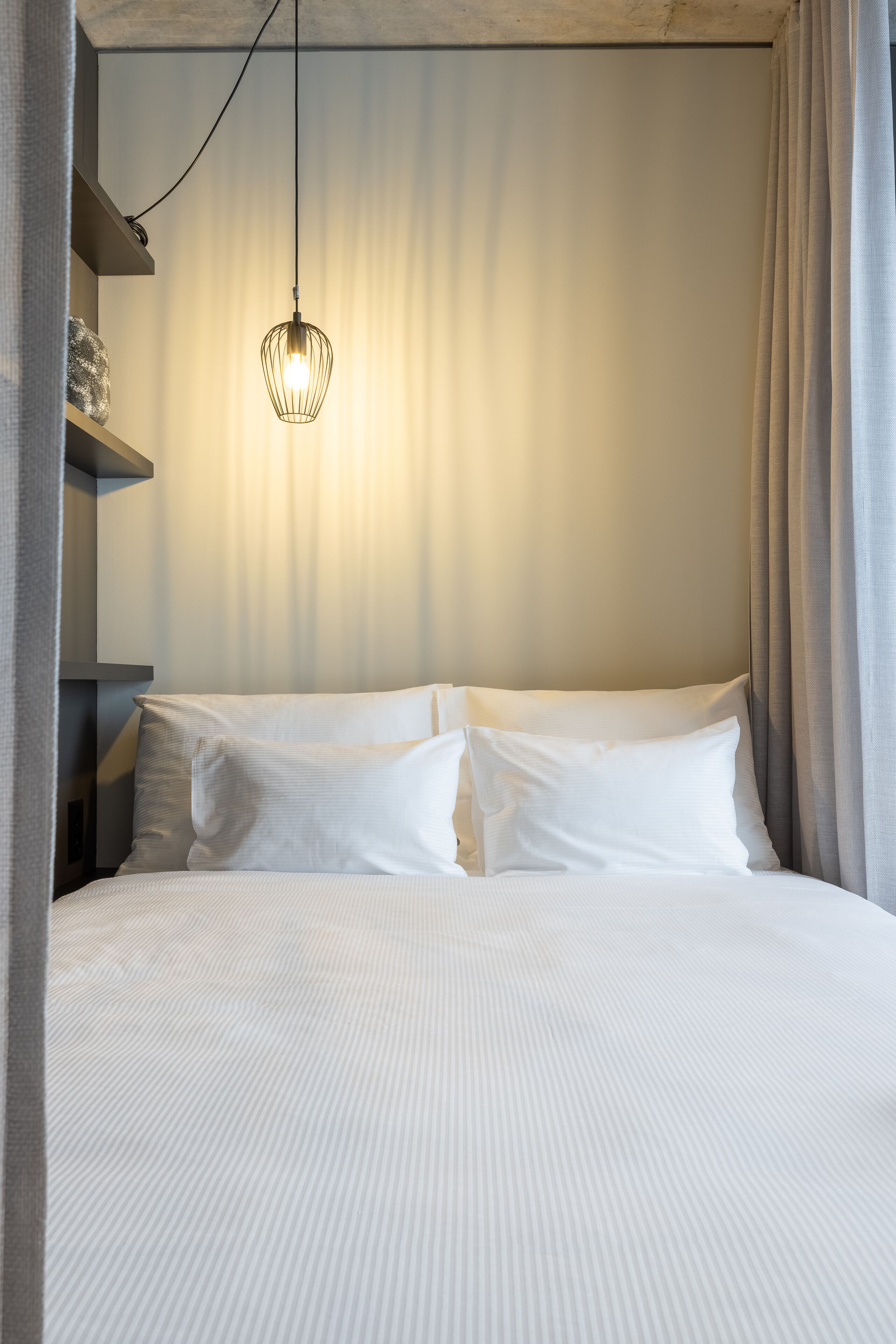 Un lit bien fait, avec du linge de lit blanc, un luminaire suspendu et un rideau à la fenêtre.
