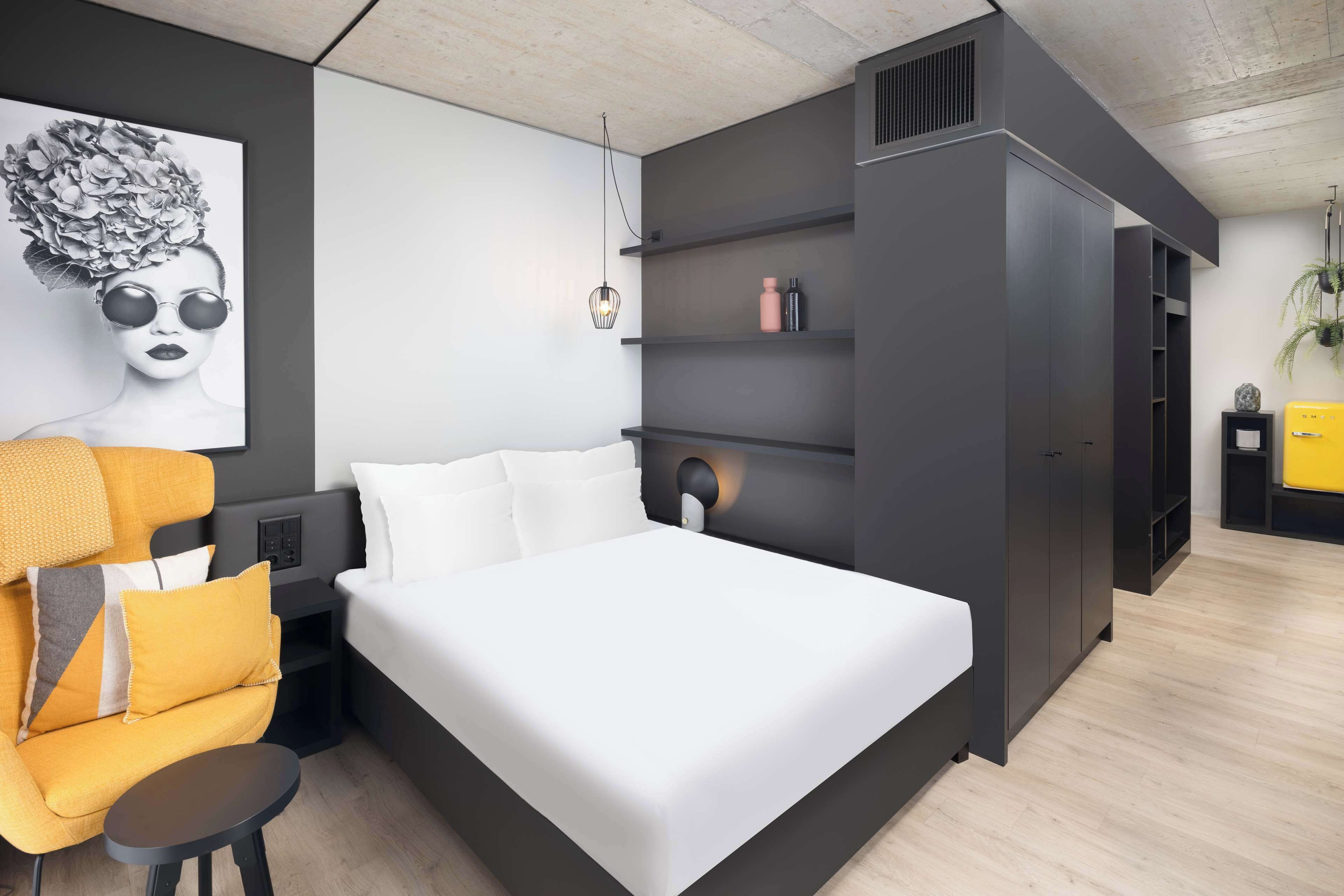 Quarto moderno com uma cama grande, um sofá, uma mesa de cabeceira, uma prateleira na parede e uma planta pendente.