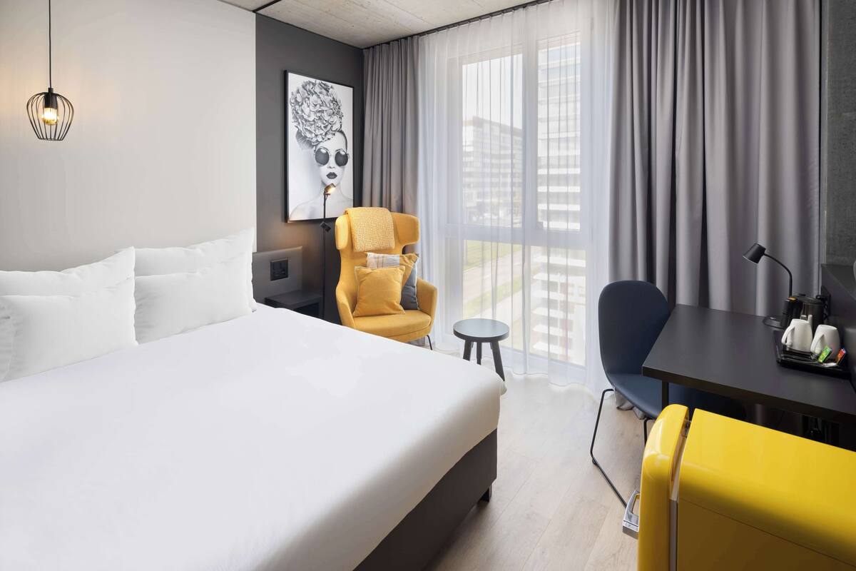 A modern hotel room with a large bed, a yellow armchair, a small round table with a lamp, and a view of the city through large windows.