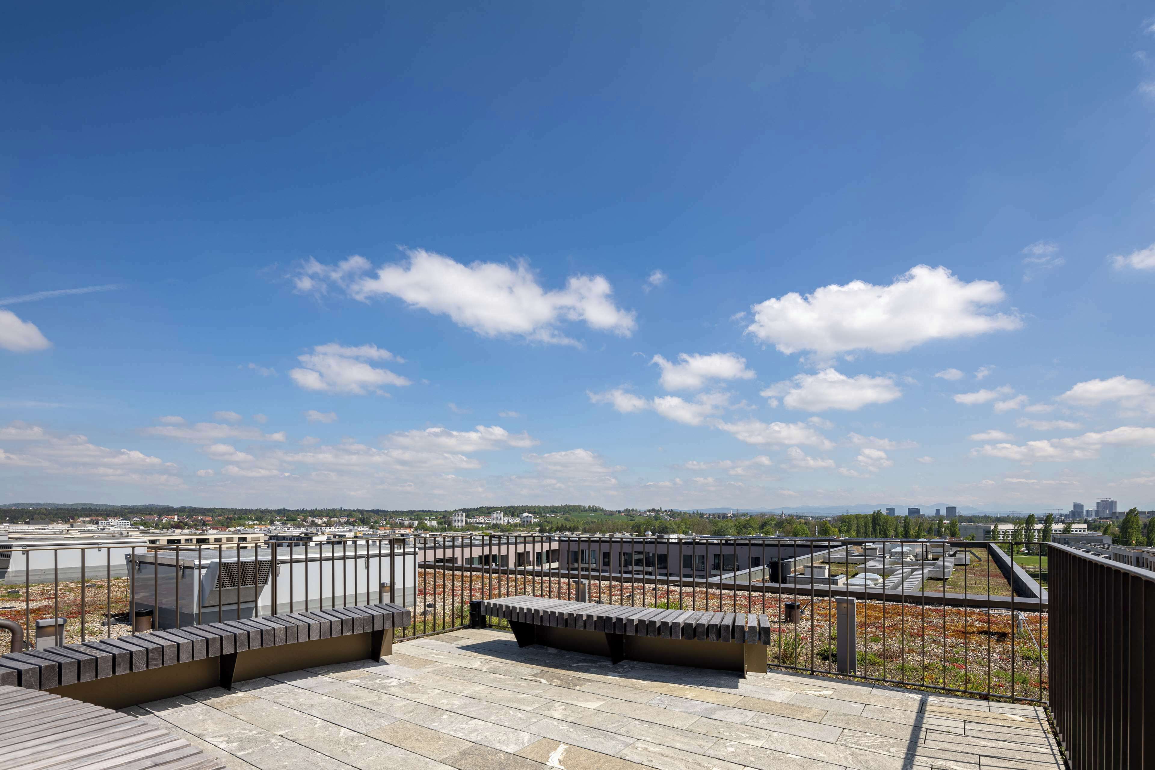 Eine Dachterrasse mit Blick auf die Stadtlandschaft, einem Metallgeländer und einer Sitzgelegenheit.