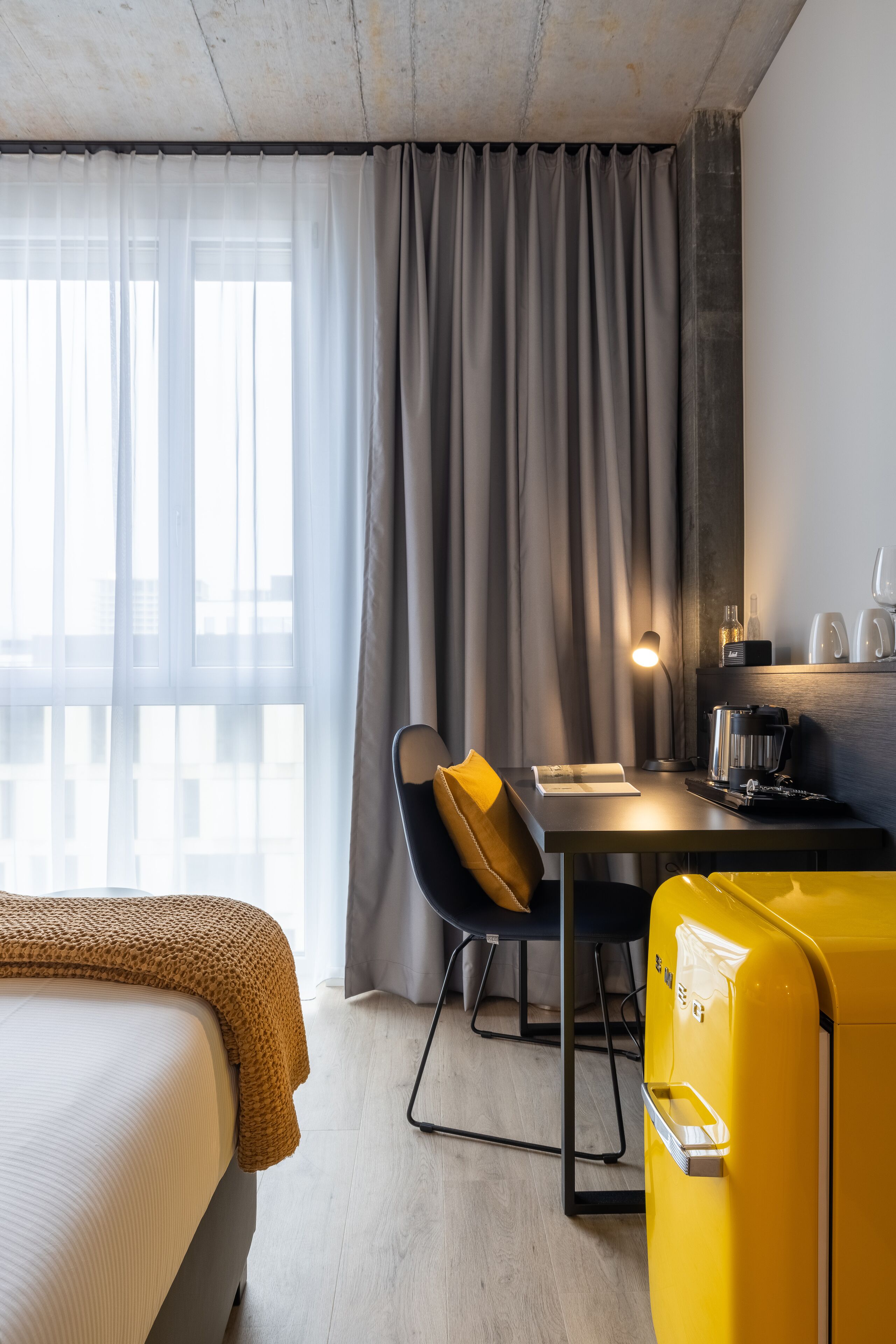 Una moderna camera d'albergo con un letto, una scrivania con una lampada, una valigia gialla e vista sulla città attraverso ampie finestre.