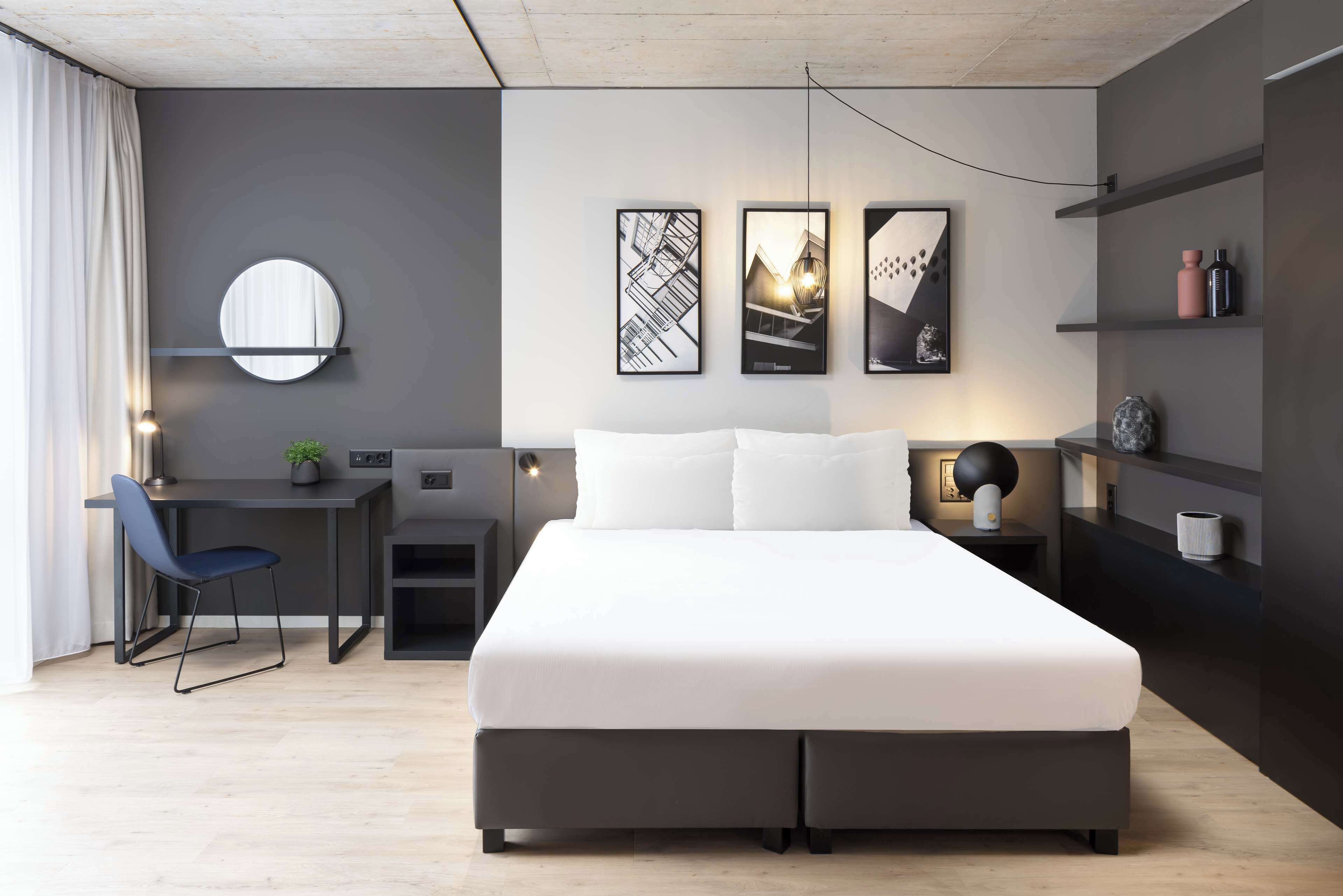 Habitación de hotel moderna con una cama grande, un escritorio y una silla. Hay tres cuadros en la pared, un espejo redondo y una estantería con objetos decorativos.
