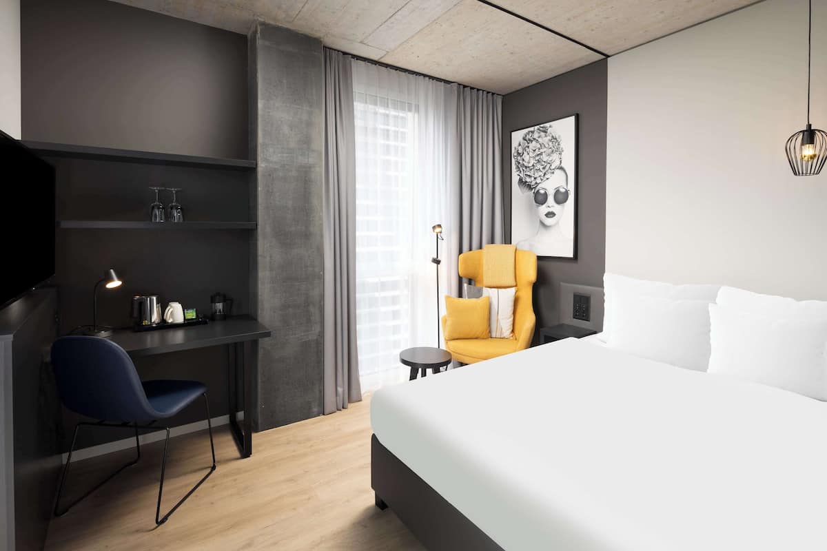 A modern hotel room with a large bed, a desk with a chair, a small table, and a chair with a yellow cushion.