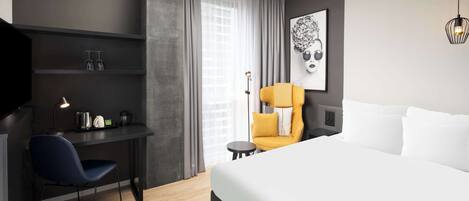 Habitación de hotel moderna con una cama grande, un escritorio con silla, una mesita pequeña y un sillón con cojín amarillo.