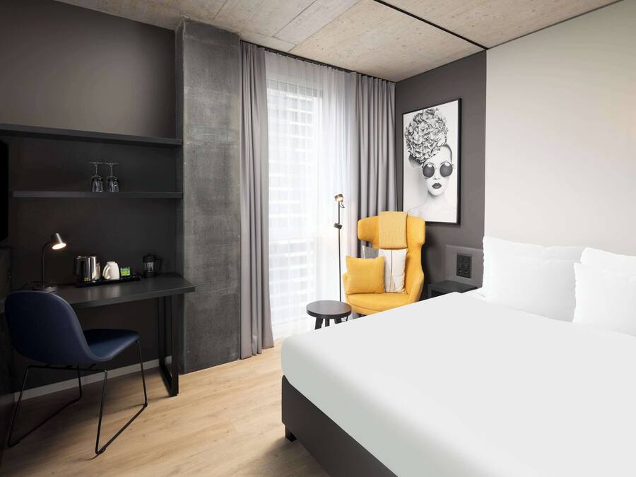 A modern hotel room with a large bed, a desk with a chair, a small table, and a chair with a yellow cushion.