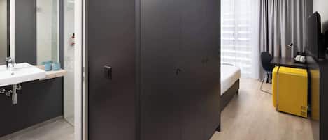 A modern hotel room with a large wardrobe, a bed, a desk with a chair, a small yellow refrigerator, and a bathroom with a sink and mirror.