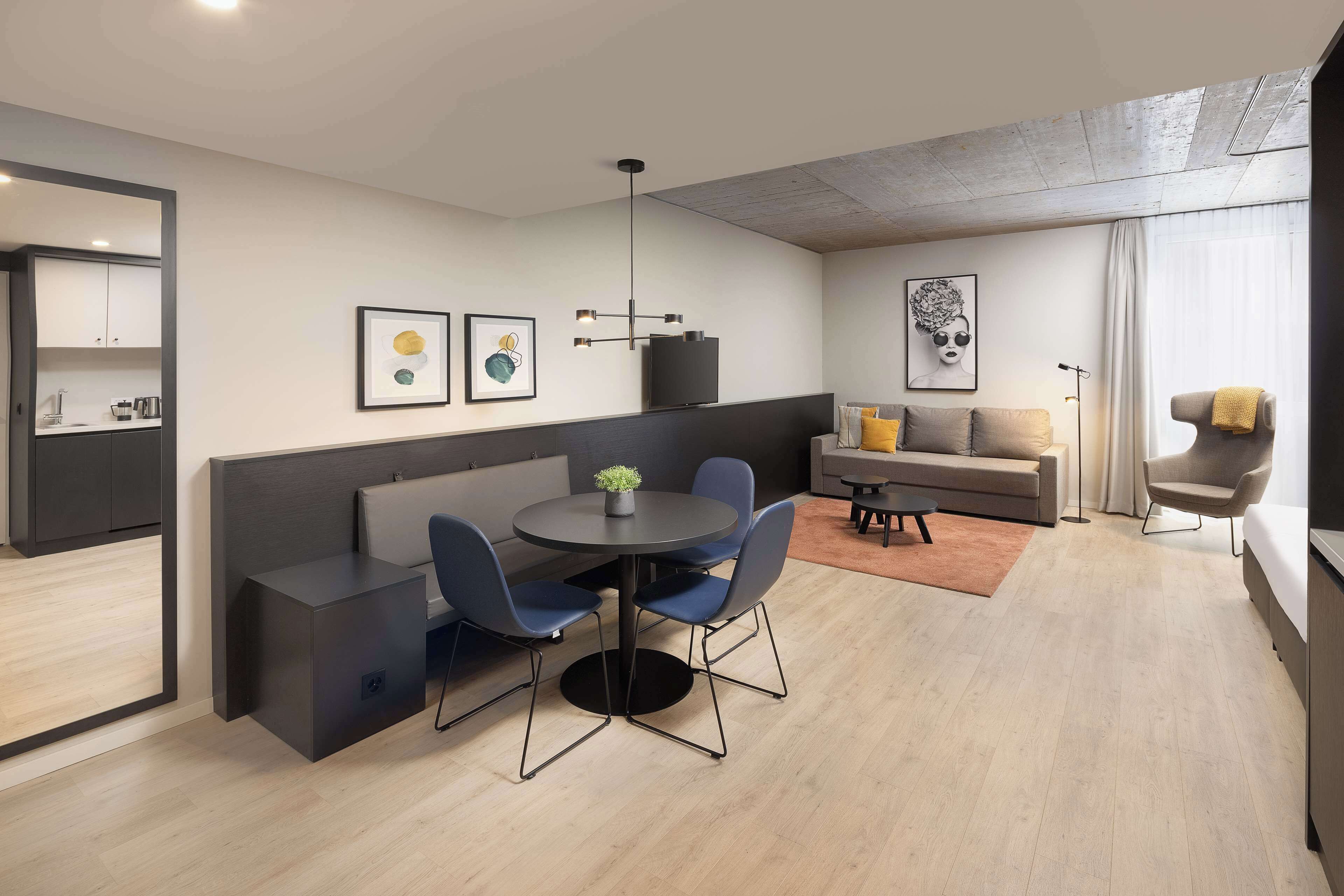 A modern living room with a round dining table, blue chairs, a sofa, and a coffee table. There is a television on a stand, a plant on the table, and artwork on the walls.
