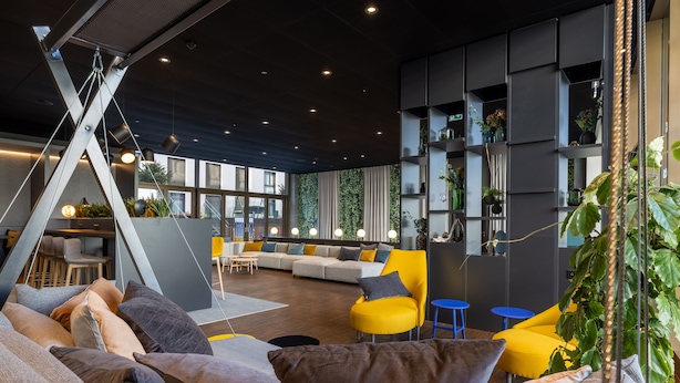 A modern lounge area with a sectional sofa, yellow chairs, and a hanging plant. - Radisson Hotel & Suites Zurich (Opfikon)