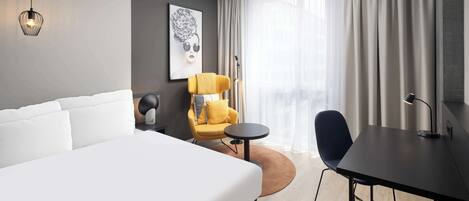 A modern hotel room with a large bed, a yellow armchair, a small round table, and a desk with a lamp.