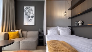 A modern hotel room with a grey sofa, a small round table, a bed with white linens, a patterned throw blanket, and a wall-mounted artwork. - Radisson Hotel & Suites Zurich (Opfikon)