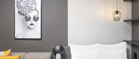 A modern bedroom with a grey sofa, a white bed, a black lamp, and a framed artwork on the wall.