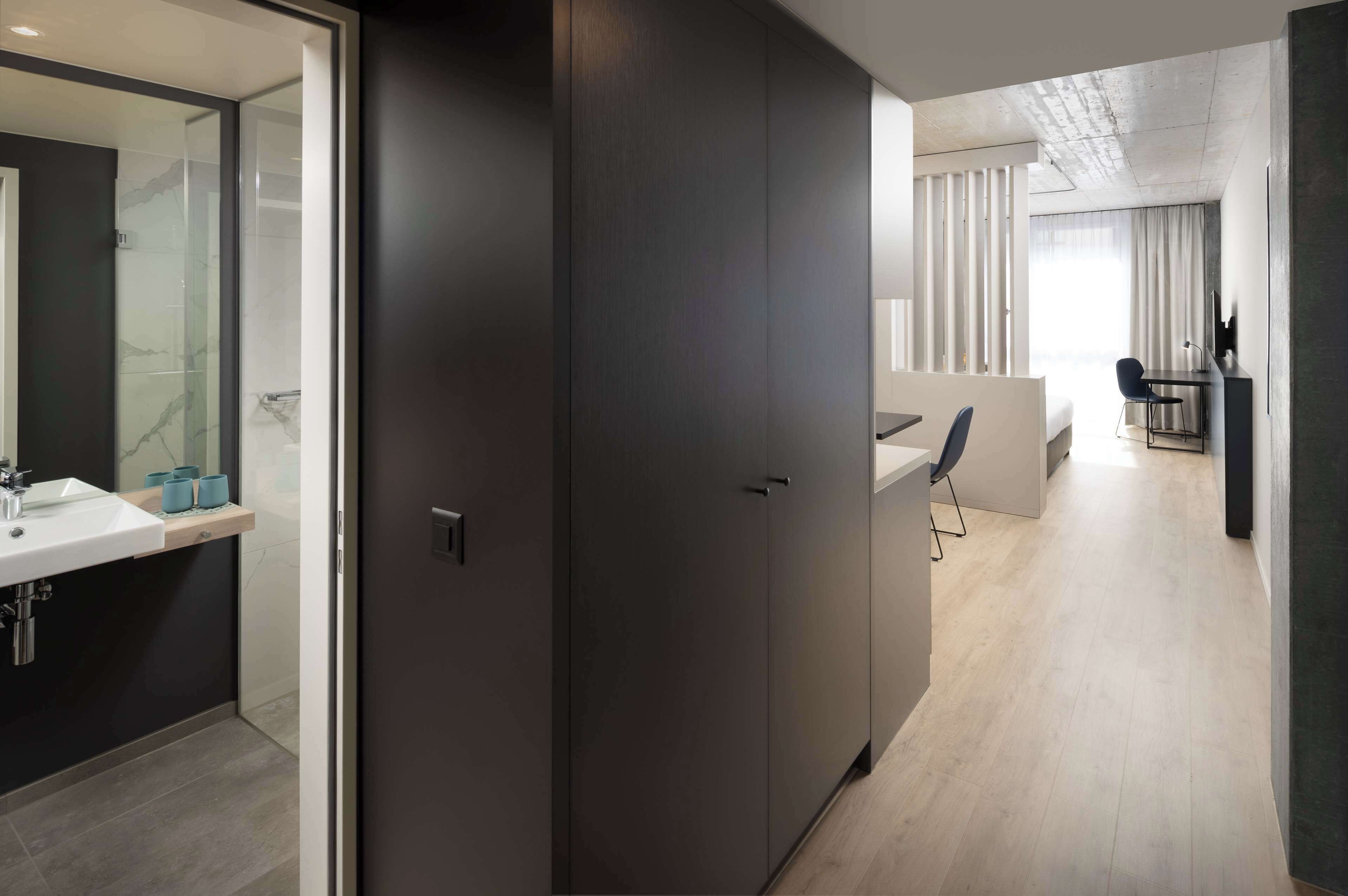 A modern hotel room with a large sliding door, a bathroom with a glass shower, a bed, and a desk area with a chair.