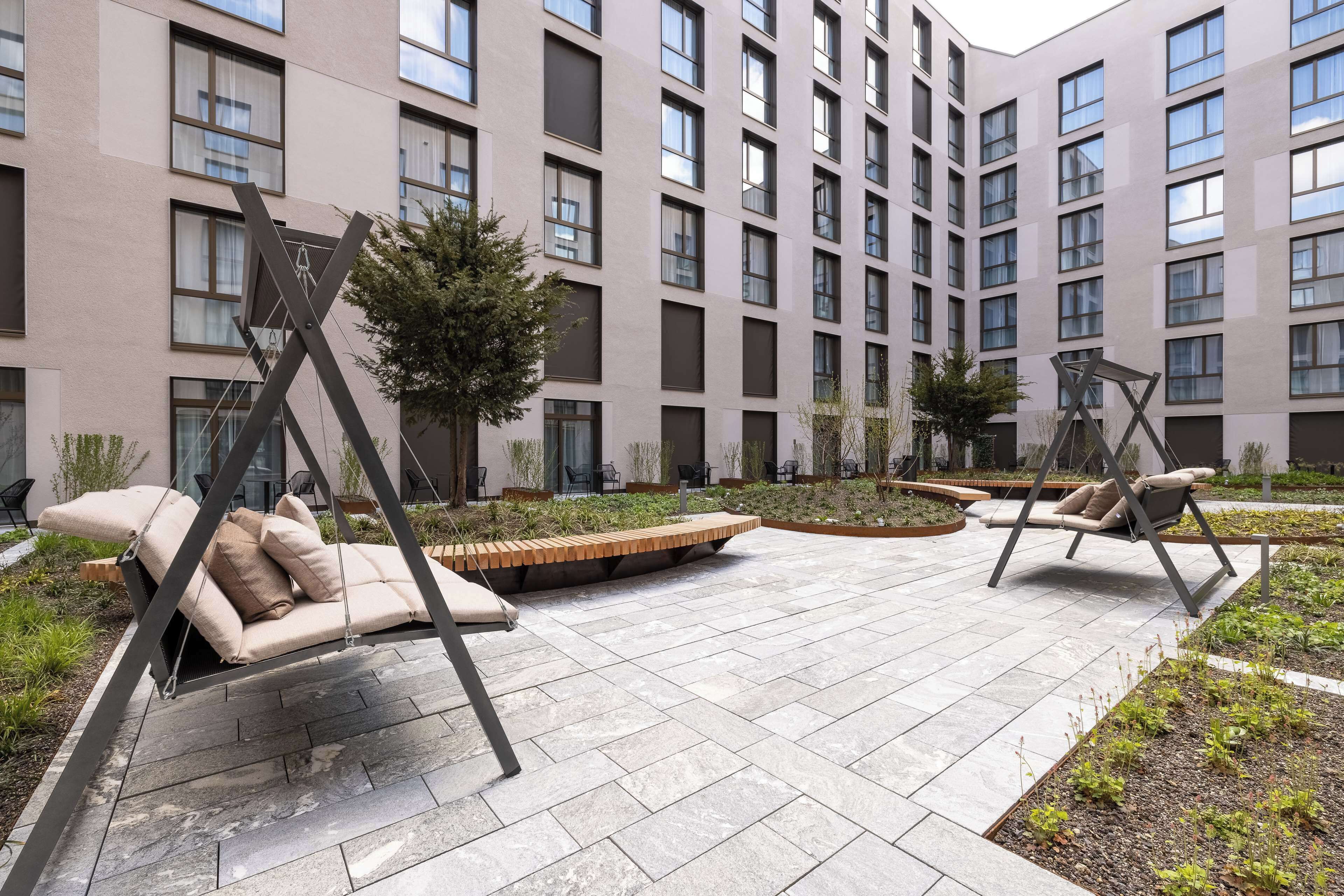 Un patio exterior moderno con sillas tipo columpio y una zona de estar, rodeado por un edificio con múltiples ventanas y un jardín ajardinado.
