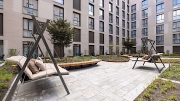Un patio exterior moderno con sillas tipo columpio y una zona de estar, rodeado por un edificio con múltiples ventanas y un jardín ajardinado.
