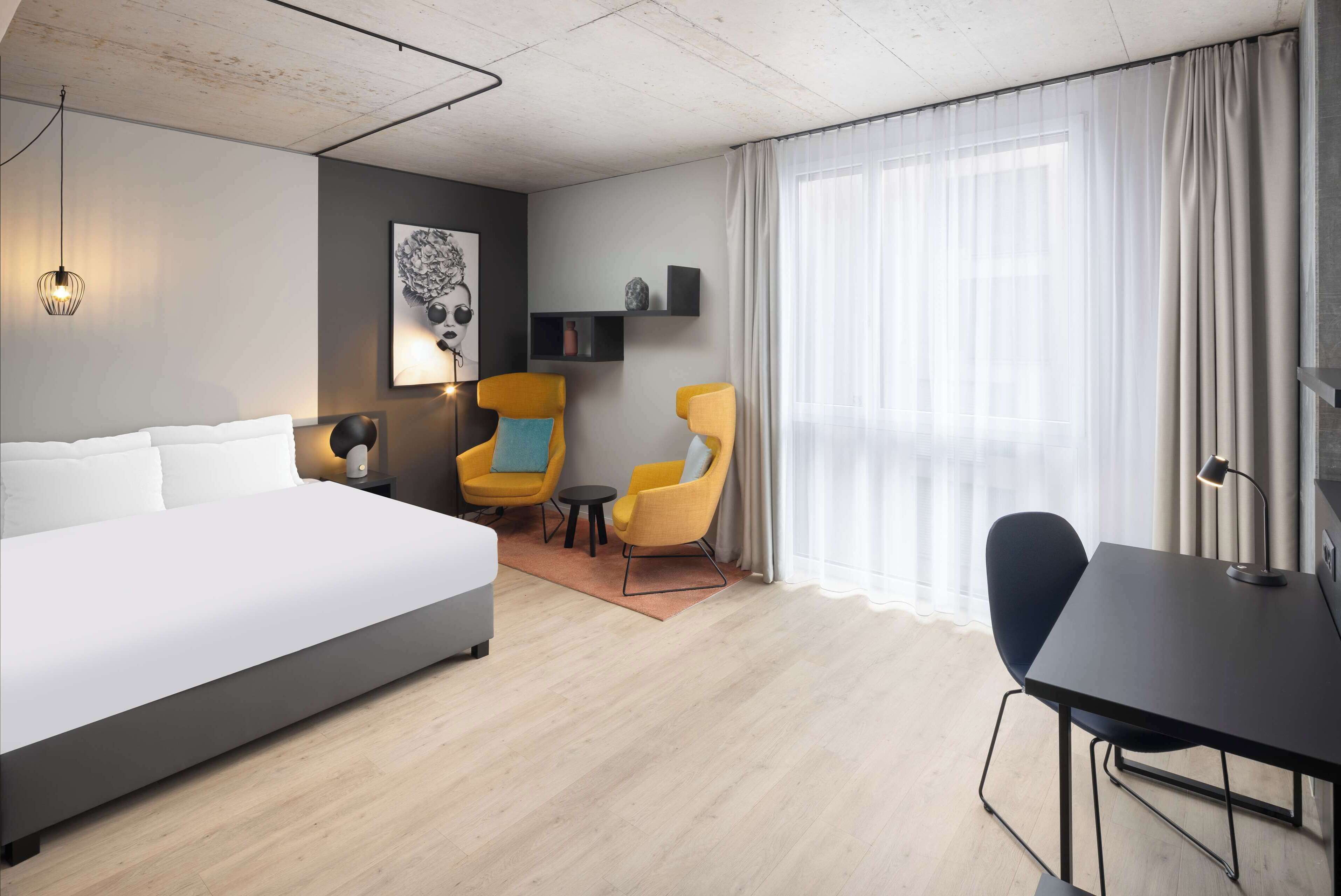 A modern hotel room with a large bed, two yellow armchairs, a small round table, a desk with a chair, and a large window with sheer curtains.