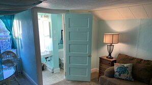 A room with a teal door, a small table with a lamp, a brown sofa with a pillow, and a chair. - Rustic Rooftop Delight - Bay Front 2 Bedroom Apts by RedAwning (Norfolk)