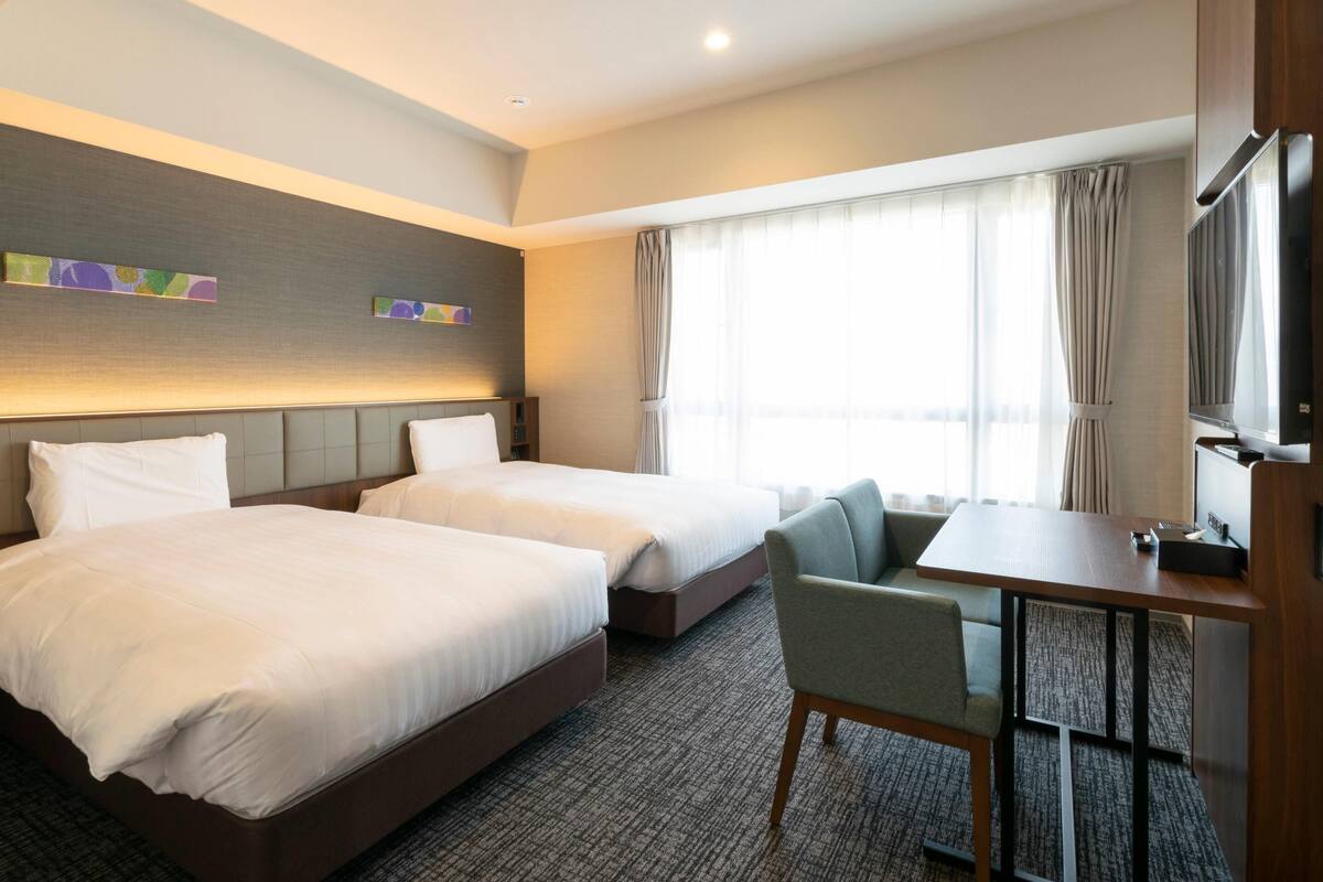 Habitación de hotel con dos camas, un escritorio, una silla, un televisor y un ventanal con cortinas.