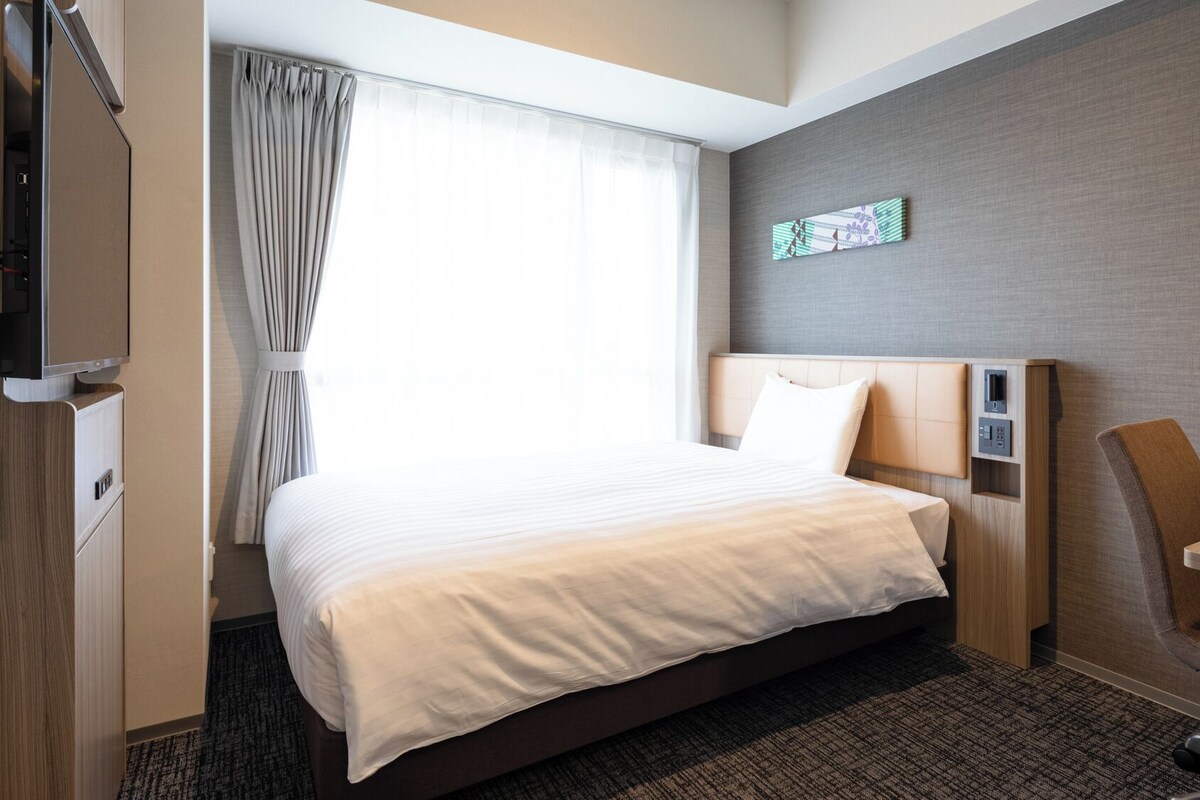 Une chambre d’hôtel équipée d’un lit, d’un bureau, d’une chaise, d’une télévision et d’une fenêtre avec des rideaux.