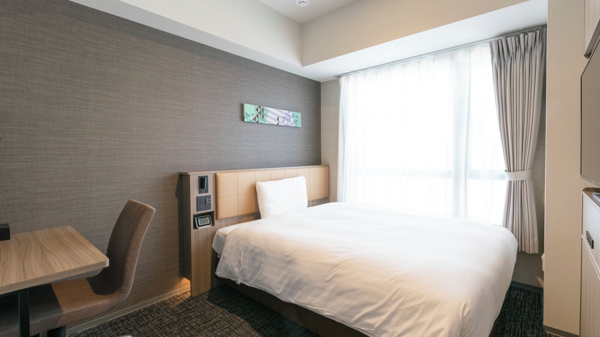 A hotel room with a bed, a desk, a chair, a small table, a wall clock, and a window with curtains.