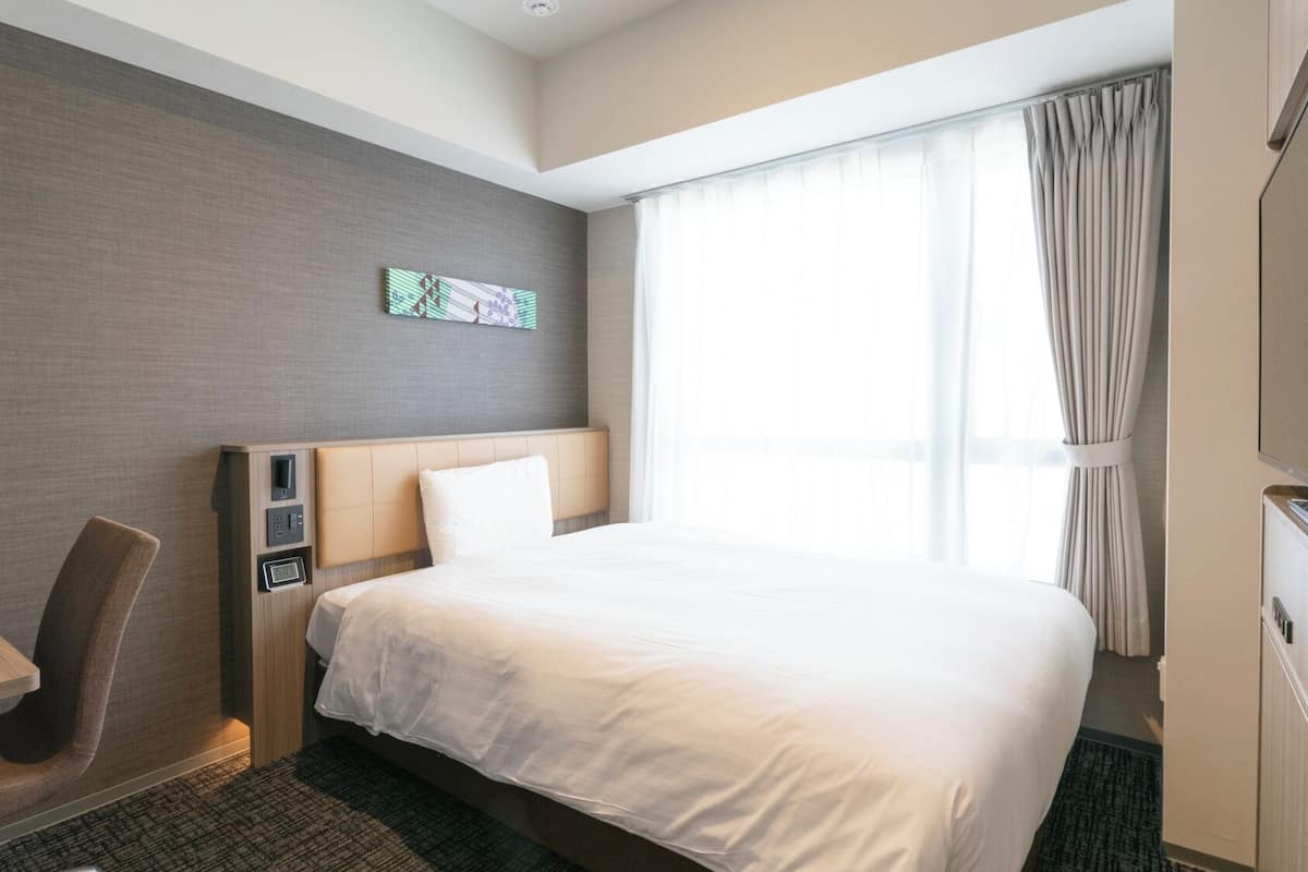 Une chambre d’hôtel comprenant un lit, un bureau, une chaise, une petite table, une horloge murale et une fenêtre avec des rideaux.