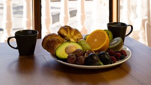 Mesa de café da manhã com croissants, frutas e uma xícara, com vista para uma varanda com corrimão e prédios ao fundo.