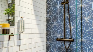 Un baño con paredes de azulejos blancos, pared de ducha con estampado geométrico azul y un estante de vidrio con artículos de aseo, además de un lavabo blanco.