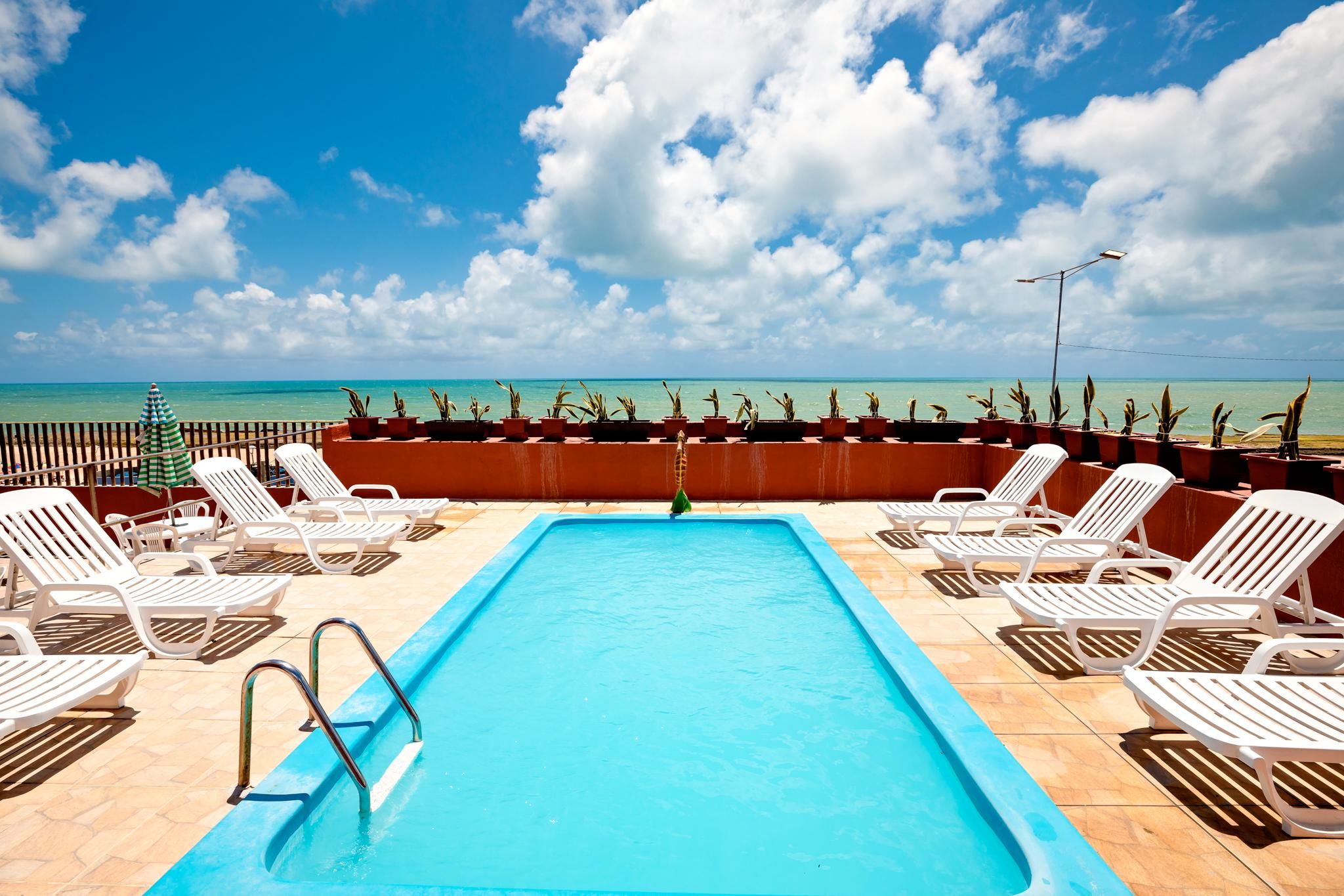 Área de piscina com espreguiçadeiras e plantas em vasos, com vista para uma praia.
