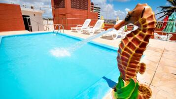 Uma piscina com uma estátua de cavalo-marinho, cadeiras de descanso brancas e um edifício de tijolos vermelhos ao fundo.