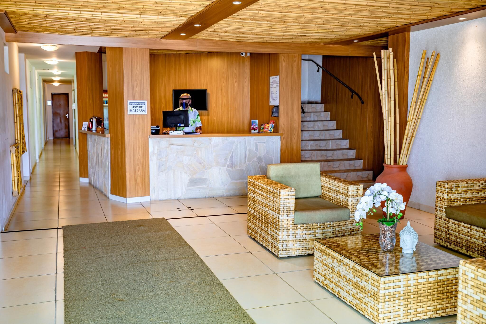 Un área de recepción con un mostrador de madera, una pantalla, una recepcionista, mobiliario de mimbre, una escalera y un adorno de bambú.
