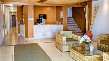 Un área de recepción con un mostrador de madera, una pantalla, una recepcionista, mobiliario de mimbre, una escalera y un adorno de bambú.