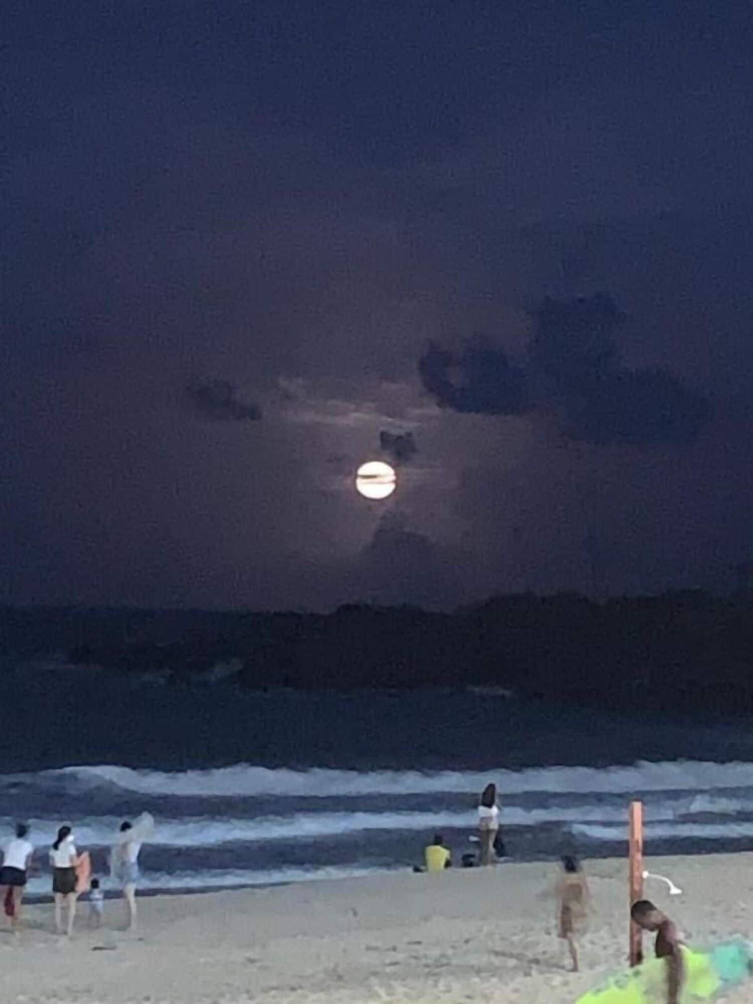 A beach scene with people and a moon in the sky.
