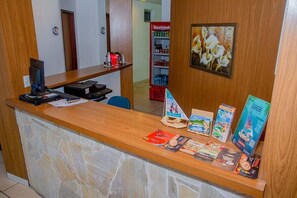 Un mostrador de recepción con una computadora, documentos y folletos. Se ve un refrigerador al fondo con bebidas.