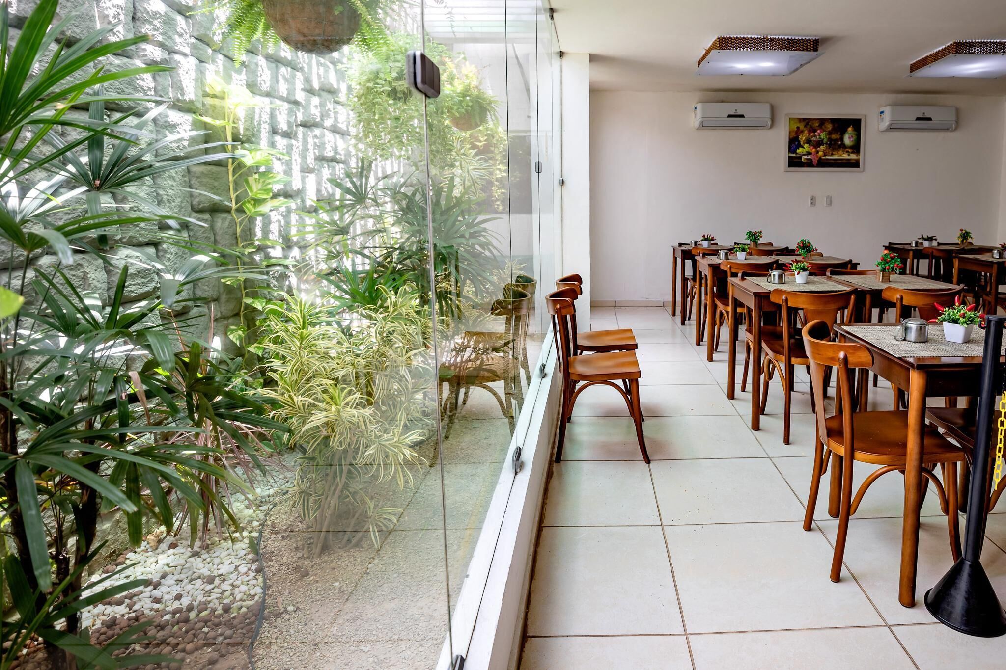 Comedor con mesas y sillas de madera, un separador de vidrio y vistas a un jardín con diversas plantas.