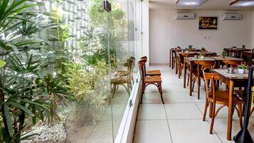 Comedor con mesas y sillas de madera, un separador de vidrio y vistas a un jardín con diversas plantas.