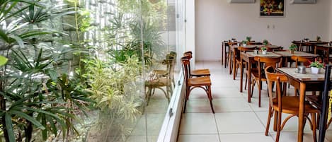 Comedor con mesas y sillas de madera, un separador de vidrio y vistas a un jardín con diversas plantas.