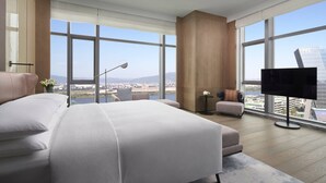 A modern hotel room with a large bed, a TV, a small table, and a view of the cityscape through large windows.