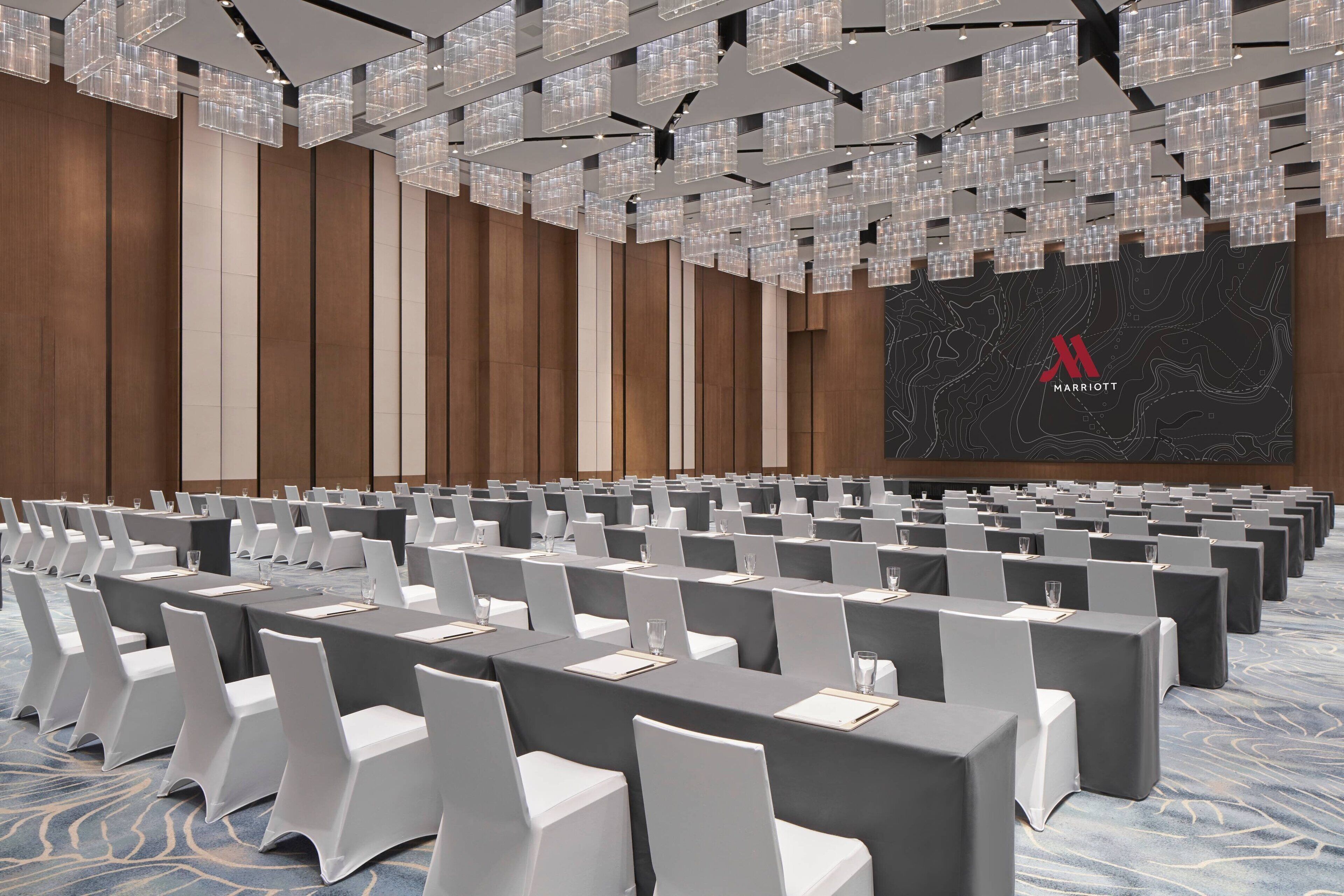 A conference room with rows of tables and chairs, a large screen with the Marriott logo, and a modern ceiling design.