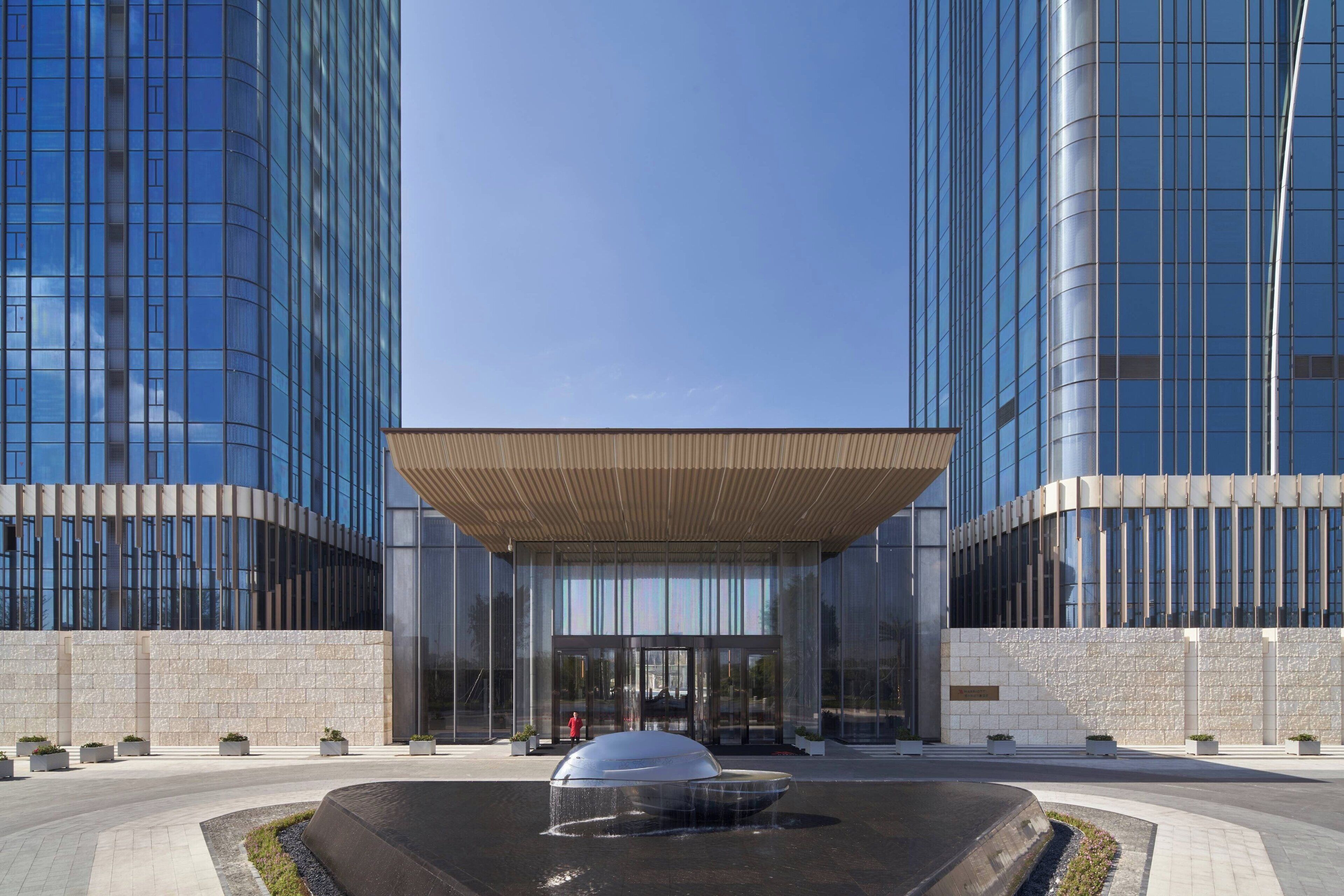 Un bâtiment moderne doté d’une façade en verre, d’une entrée centrale surmontée d’un auvent et d’un élément décoratif aquatique à l’avant.