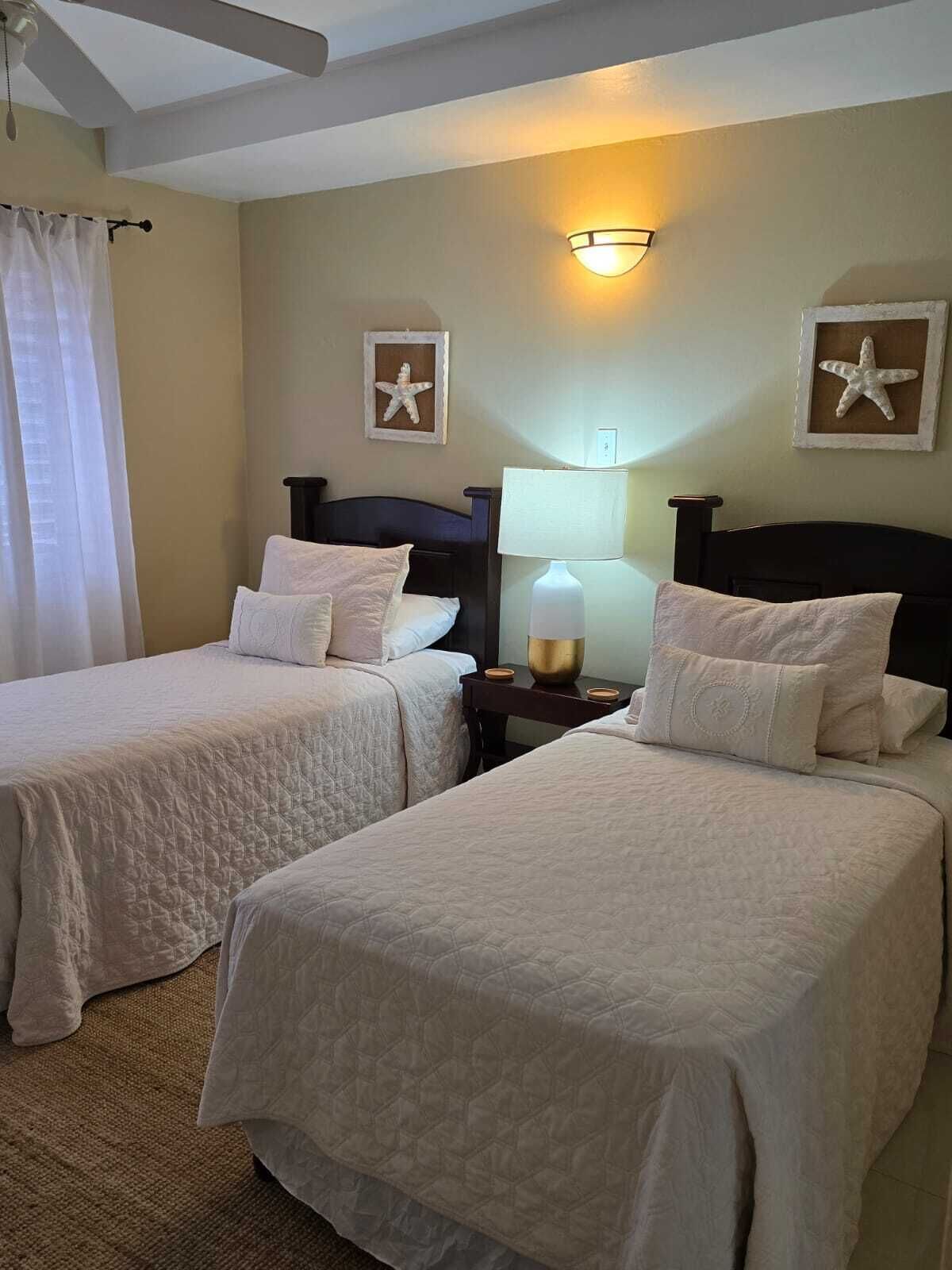 A hotel room with two beds, a nightstand with a lamp, starfish wall art, and a ceiling fan.