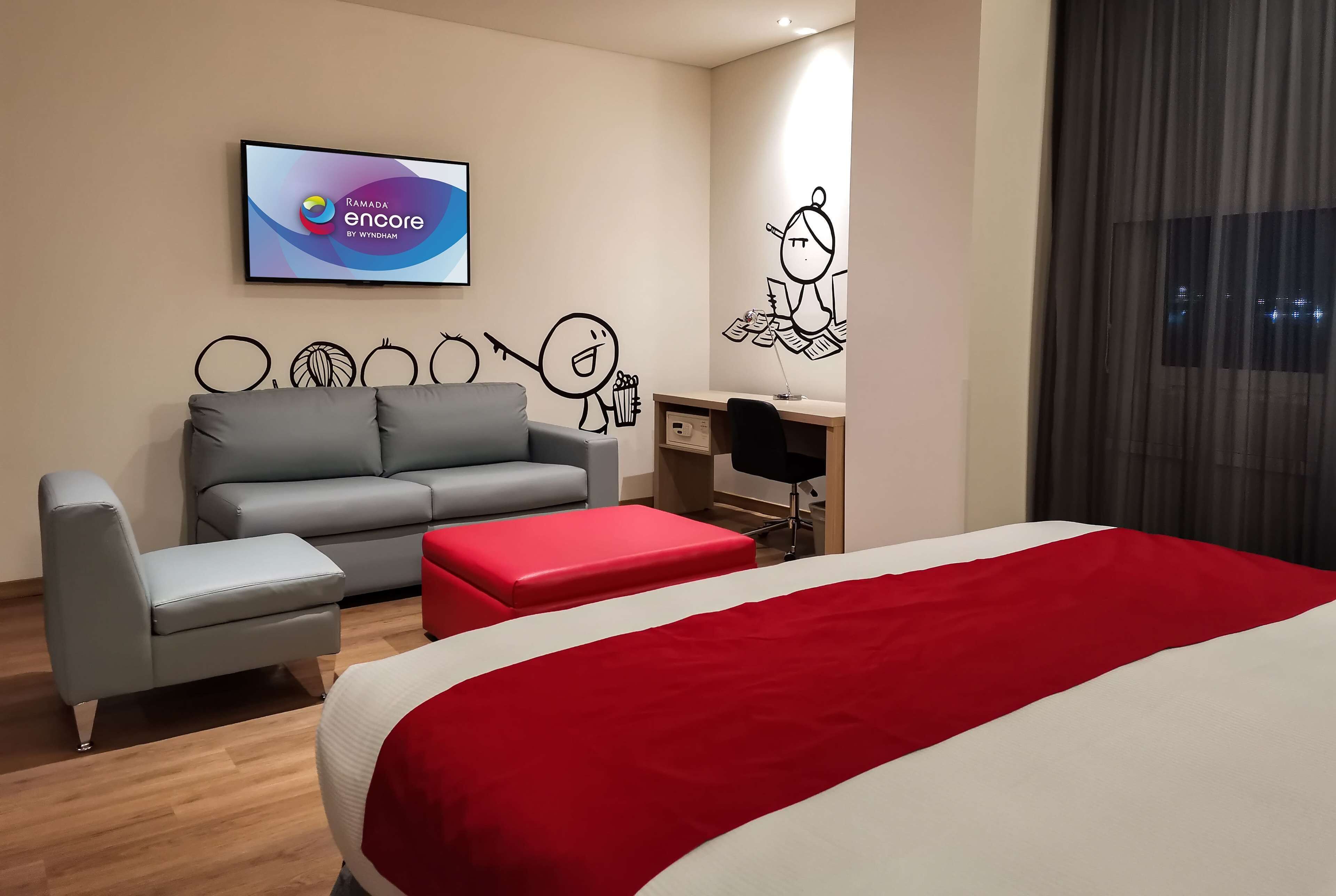 A modern hotel room with a bed, grey sofa, red ottoman, and a desk with a chair.