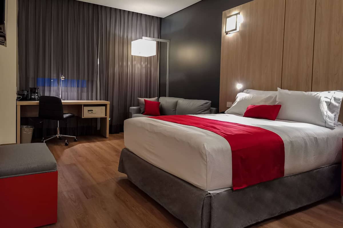 A modern hotel room with a bed, desk, chair, and a red accent piece.