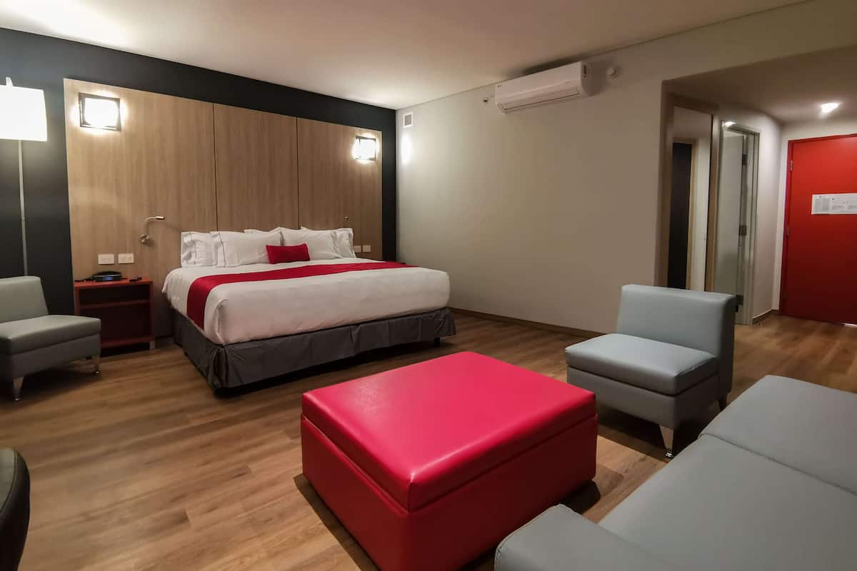 A modern hotel room with a wooden headboard, a large bed with white bedding and red accents, a red ottoman, grey armchairs, and a red door.