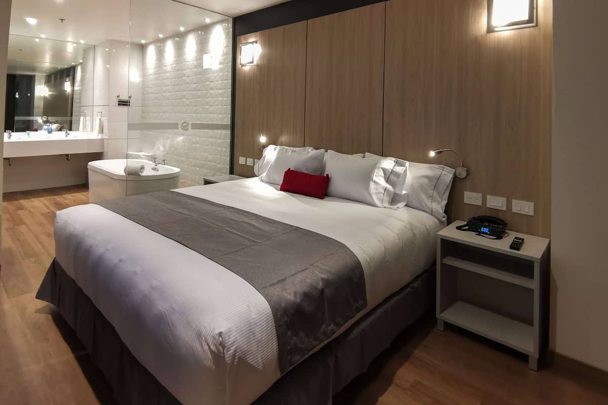A modern hotel room with a large bed, a bedside table with a phone, a nightstand, a bathroom with a sink and mirror, and a wooden wall paneling.
