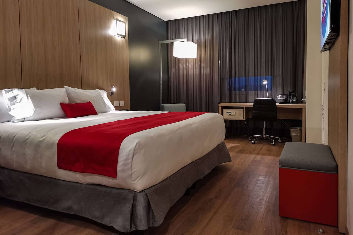 A hotel room with a bed, desk, chair, and a red ottoman.